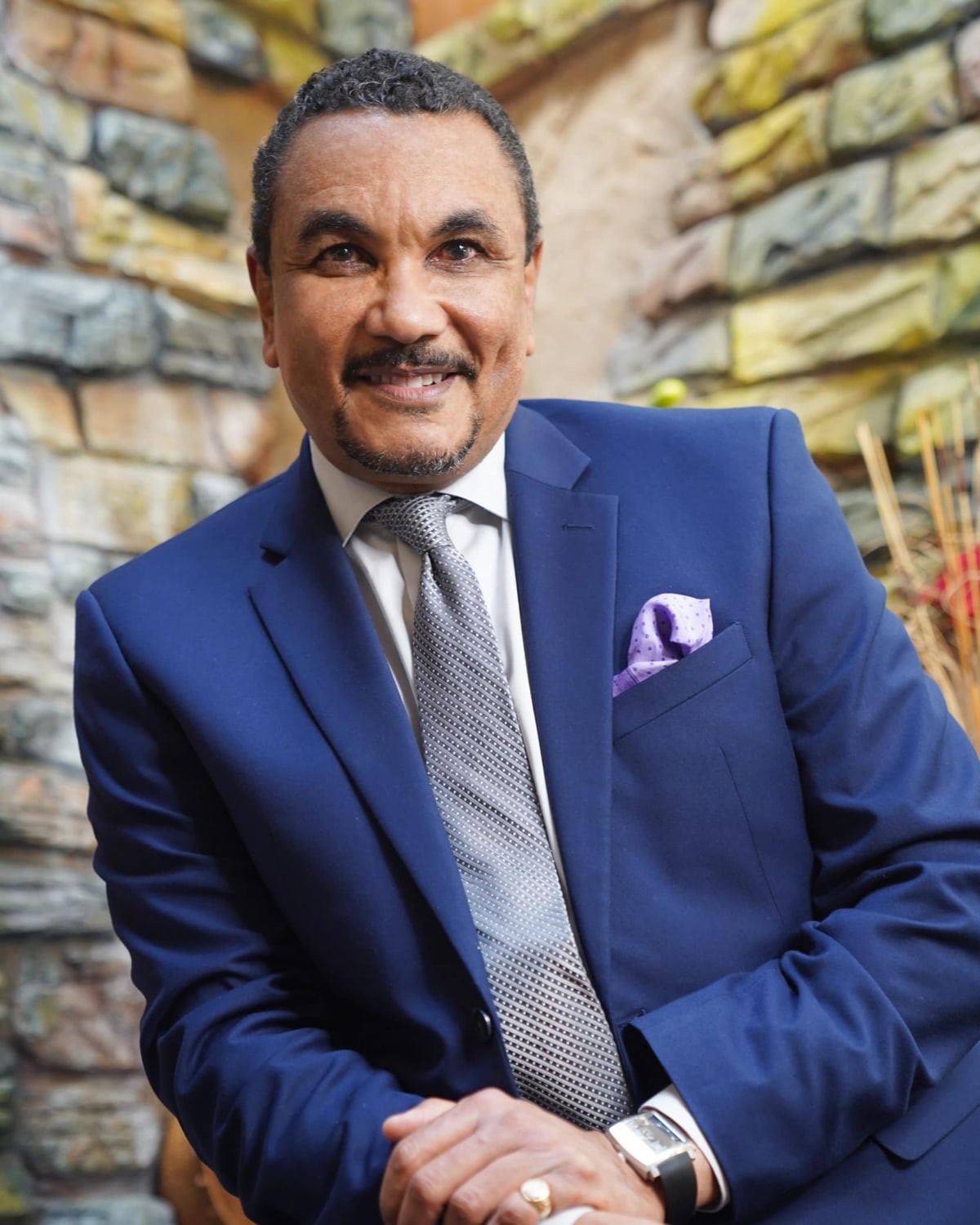 Dr. Kenrick Spence in a blue suit and silver tie, smiling, seated before a stone wall