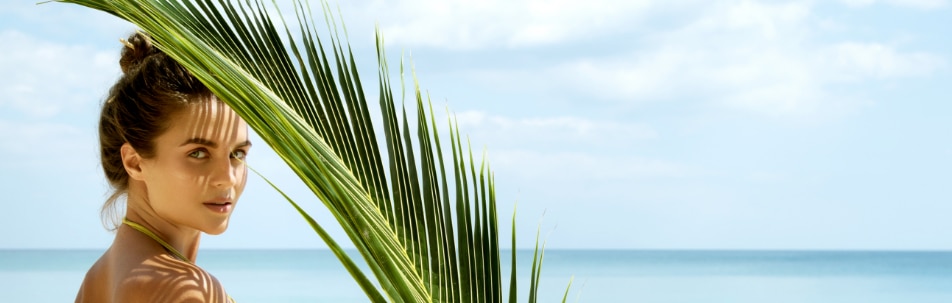 A woman looking back over her shoulder beside a palm frond, with the sea behind her