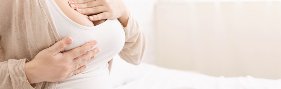 Woman in a white top sitting on a bed with her hands on her breasts