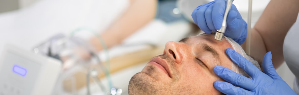 Man with eyes closed while a gloved technician uses a microdermabrasion wand on his forehead